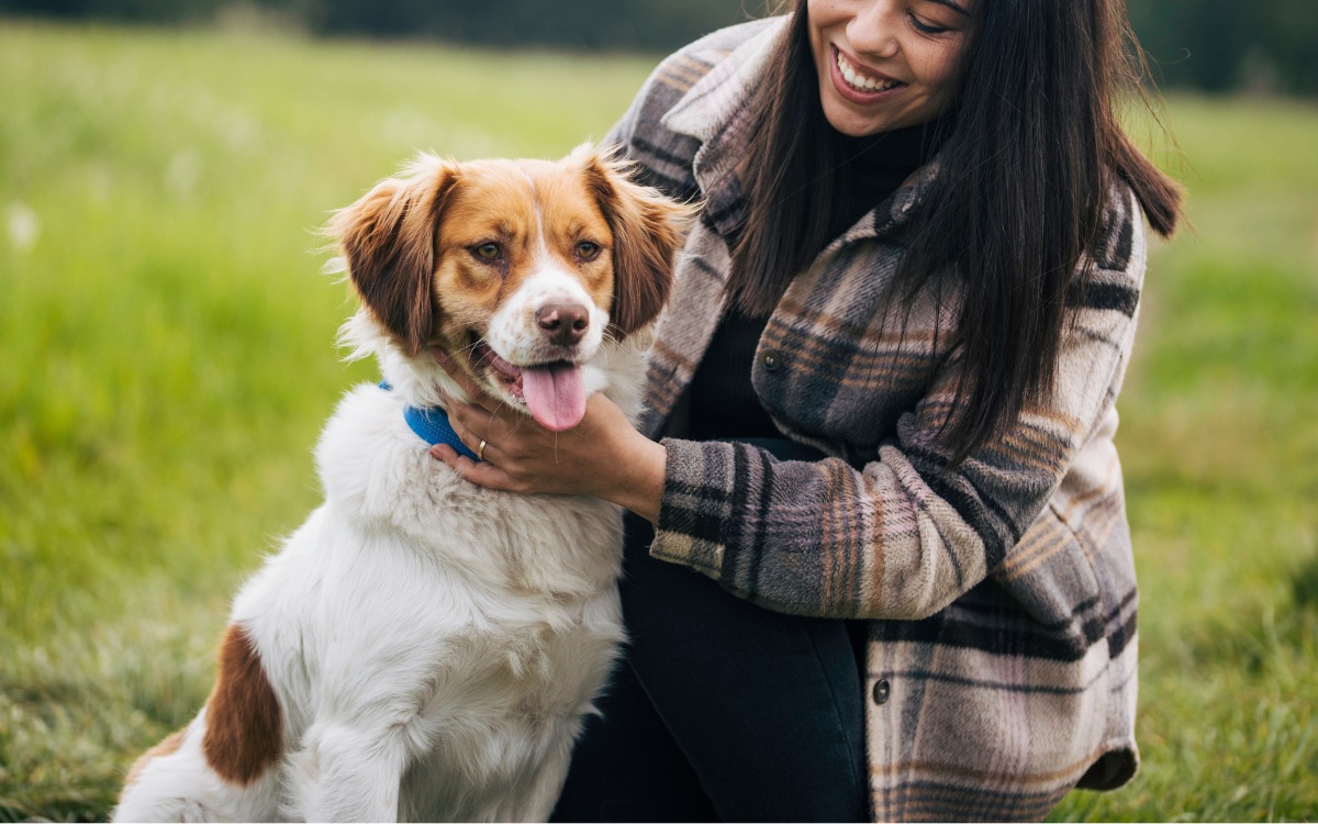 A woman gently pets her dog while standing in a lush green field